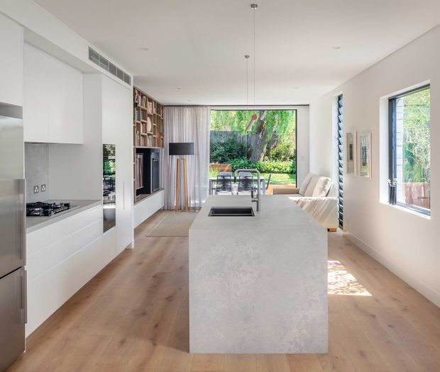A bright, open-plan contemporary kitchen and living area with a large pale stone island, white handleless cabinetry, integrated appliances, timber flooring, and a built-in media wall with shelving. The island looks toward a dining area and glazed garden-facing opening, while side windows bring in abundant natural light.