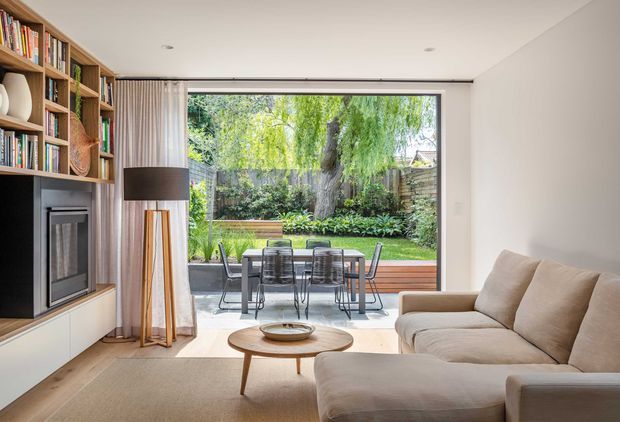 Bright contemporary living room with a built-in timber bookcase and fireplace along the left wall, a large cream sectional sofa, and a round timber coffee table. Full-width glazing opens the room to a timber deck with outdoor dining and a leafy garden beyond. The space has a clean, warm, refined character with integrated storage and a low, minimalist ceiling.