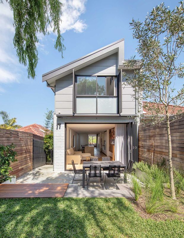 A contemporary two-storey residential extension or compact house viewed from the rear garden. The facade combines horizontal light-grey cladding, dark-framed glazing and a sharply angled skillion roof. Large glazed folding or sliding doors open the ground-floor living area onto a paved patio with an outdoor dining setting, timber decking and landscaped lawn. Timber privacy fences and established planting frame the courtyard-like backyard.