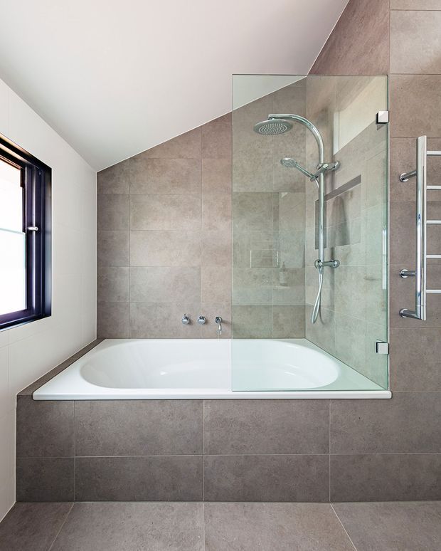 Modern bathroom featuring a built-in white bathtub with an overhead and handheld shower behind a frameless glass screen. Large matte taupe-grey stone-look tiles cover the bath surround and walls, with matching floor tiles. A dark-framed window provides natural light, and a chrome heated towel rail is mounted beside the shower.