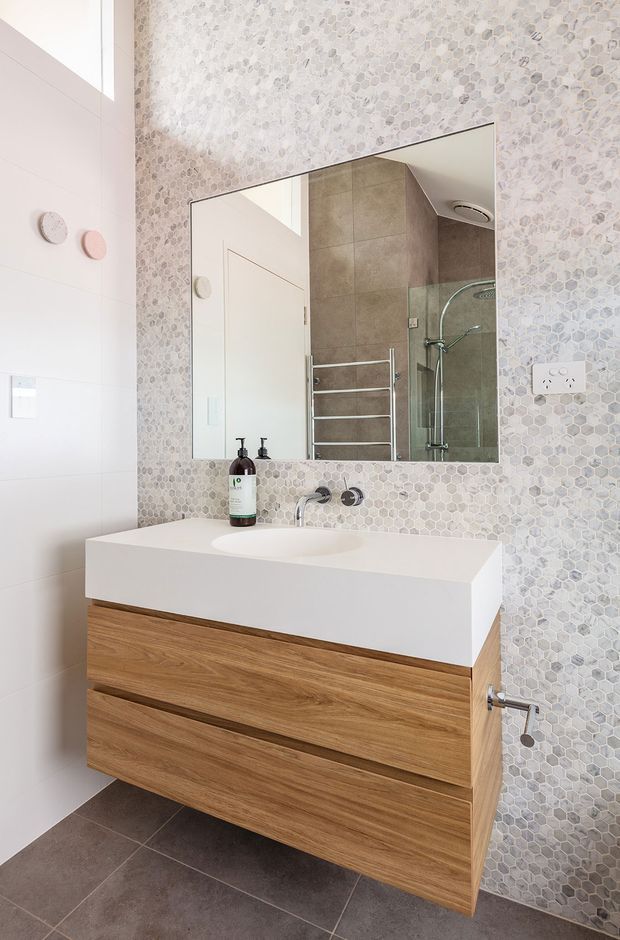 Bright contemporary bathroom vanity with a wall-mounted natural timber drawer unit, white integrated basin and benchtop, frameless mirror, and wall-mounted tapware. The walls feature small pale grey and white hexagonal mosaic tiles, while the floor is finished in large dark grey tiles. A glass-enclosed shower and heated towel rail are visible in the mirror reflection.