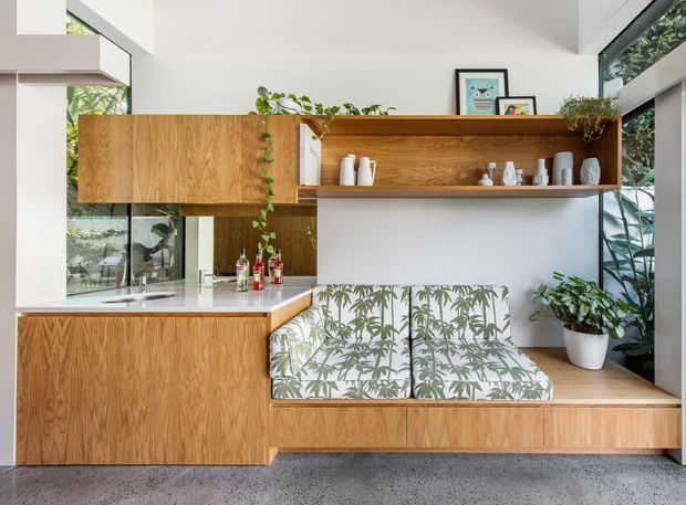A bright, contemporary open-plan kitchen and built-in dining nook. The space features warm timber cabinetry and shelving, a pale stone benchtop, white walls, expansive glazing, indoor plants, and a built-in timber bench with botanical-patterned cushions. Decorative ceramics and artwork add a relaxed, nature-inspired character.