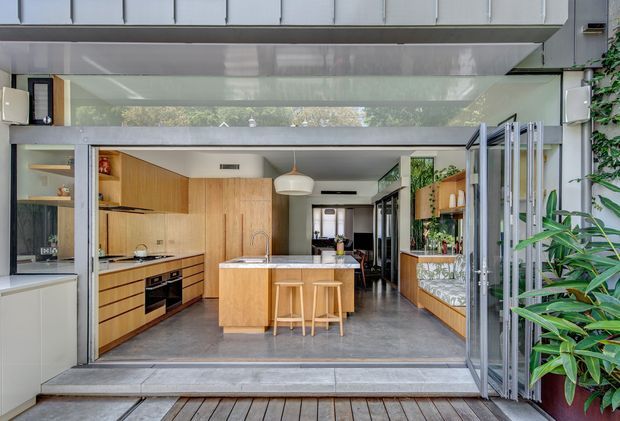 A bright, contemporary kitchen viewed from an outdoor deck through fully opened glazed folding doors. The room features extensive light timber cabinetry, a central island with bar stools, integrated appliances, a grey polished floor, a white ceiling with pendant lighting, and indoor planting. The open connection to the deck creates an airy indoor-outdoor layout.