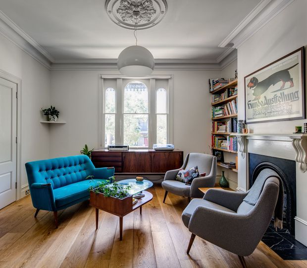 A refined living room with a period-inspired interior, featuring ornate ceiling cornice and decorative ceiling medallion, white paneled walls, a carved fireplace mantel, tall arched windows, and warm timber flooring. The room is furnished with a vivid teal tufted sofa, two grey upholstered lounge chairs, mid-century-style timber coffee tables, built-in shelving filled with books, and a dark timber sideboard. The overall atmosphere is bright, sophisticated, and eclectic, blending traditional architectural detailing with contemporary and mid-century furnishings.