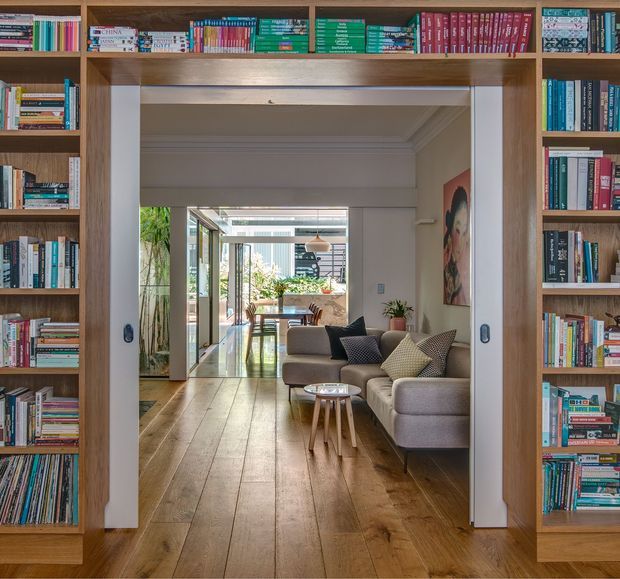 View through a wide sliding doorway framed by floor-to-ceiling built-in bookcases, looking into a warm, light-filled living room and dining area. The room has broad timber floorboards, a neutral upholstered sofa and armchair, a small nesting coffee table, indoor plants, artwork, and glazed doors opening toward a planted outdoor area. The full-height timber shelving and concealed sliding doors create a strong library-like architectural feature.
