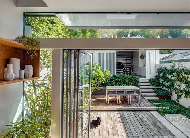 A bright indoor-outdoor transition space opening onto a landscaped courtyard and timber deck. Full-height glazing and folding glass doors connect the interior to an outdoor dining area, with built-in timber shelving and decorative objects along the left wall. The courtyard includes raised planting beds, steps, pale paving, and a white-clad structure with a visible garage beyond.