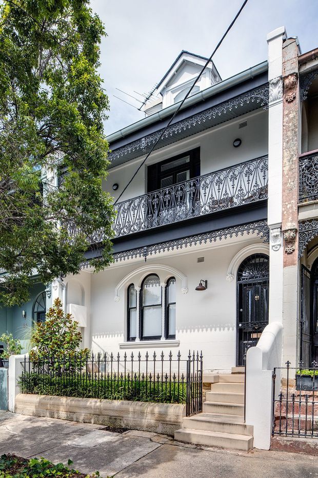 Street-level exterior view of a narrow multi-storey terrace-style residence with an ornate white-painted facade. The building features decorative wrought-iron balcony balustrades and fencing, arched windows and doorway, black entry doors, elaborate lace-like eaves detailing, masonry pilasters, and a small planted front garden behind the fence. A neighbouring attached facade is partially visible on the right, with overhead utility wiring crossing the view.