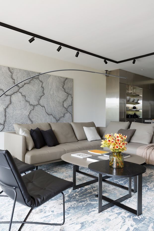 A contemporary open-plan living room with a large beige sectional sofa, black leather lounge chair, nesting coffee tables, and a patterned blue-grey area rug. A large abstract textured artwork spans the rear wall, while a compact kitchen with dark cabinetry and open shelving is visible behind the seating area. Black track lighting and a curved arc floor lamp add graphic accents to the neutral interior.