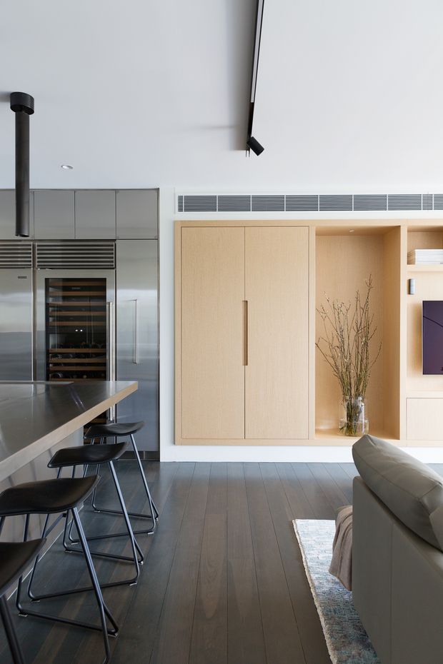 A refined open-plan kitchen and living area with a dark stone island and bar stools in the foreground, integrated stainless-steel cabinetry and wine storage to the left, and a pale timber built-in entertainment and storage wall to the right. The room has dark timber flooring, a high white ceiling with linear track lighting, concealed air-conditioning vents, and a neutral contemporary palette.