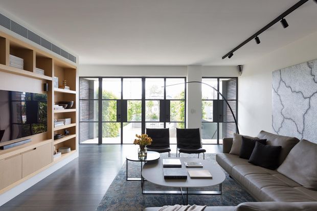 Bright contemporary living room with a large grey sectional sofa, black lounge chairs, nested oval coffee tables, and a textured area rug. Full-height black-framed glazing and louvred panels open to an exterior view, while a built-in light timber media wall with open shelving spans the left side. Black track lighting and a large monochromatic abstract wall artwork add graphic accents.