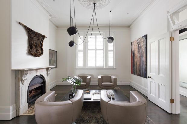 Refined formal living room with high ceilings, ornate cornice detailing and ceiling medallion, a decorative carved fireplace surround, tall shuttered window, contemporary sculptural pendant lighting, four rounded lounge chairs, and a low rectangular coffee table. The room combines classic architectural features with restrained modern furnishings and artwork in a neutral palette.