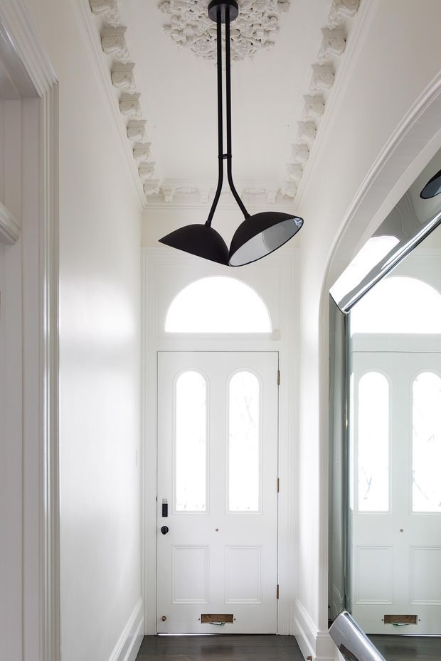 Bright, narrow residential entryway with a white paneled front door featuring arched glazing and sidelights, ornate decorative plaster ceiling mouldings, white walls and trim, a dark timber floor, and a sculptural black pendant light. A large mirrored wall reflects the door and pendant, emphasizing the corridor's height and symmetry.