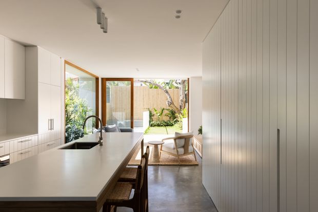 Bright open-plan kitchen opening onto a small outdoor courtyard or garden through large glazed doors. The space features a long pale stone island with integrated sink and dark timber base, white flat-panel cabinetry, full-height vertical-panel joinery, timber-framed glazing, polished concrete flooring, and a compact dining or sitting area beyond.