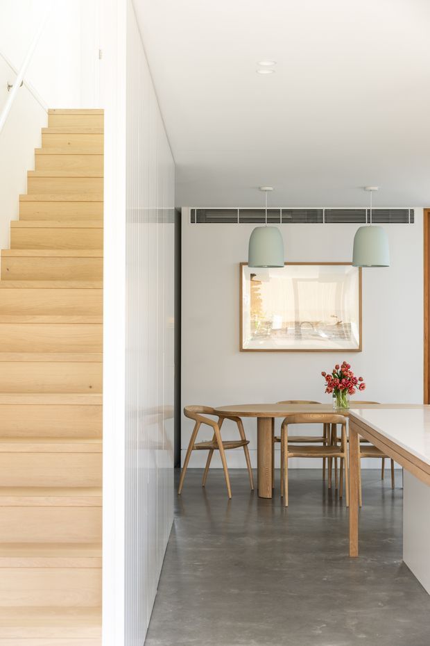 Bright contemporary dining area beside a straight timber staircase. The room features a polished grey concrete floor, pale grey walls, a white ceiling with recessed lights and linear air-conditioning grille, two muted sage pendant lights, a timber dining table with matching chairs, and a white kitchen island or bench in the foreground. A framed artwork and vase of flowers add detail.