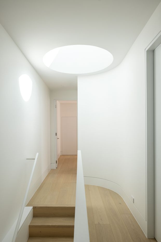 Bright minimalist interior hallway and stair landing with pale timber flooring, white walls, a curved balustrade, and a large circular ceiling skylight or light well. A partially open doorway leads to another room, while clean flush doors and concealed detailing create a seamless architectural appearance.