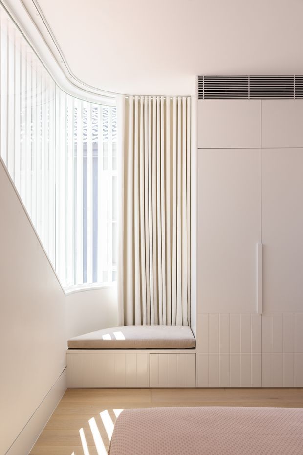 Bright minimalist bedroom corner with a curved floor-to-ceiling window, sheer vertical curtains, integrated window seat with upholstered cushion, and full-height pale built-in cabinetry. Light timber flooring and soft neutral finishes create a clean, airy interior.
