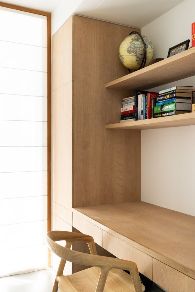 A bright built-in timber study or home office nook beside a large translucent window. Full-height warm wood cabinetry and panelled joinery frame open shelves holding books and a globe, with a broad timber desk surface and drawers below. A sculptural timber chair sits in front of the workspace. The room has white walls and ceiling, natural light, and a simple custom-built design.