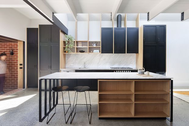 Bright contemporary kitchen with a large island featuring a light stone benchtop, dark cabinetry, open timber shelving, and a timber display/storage unit at the seating end. The room has a high white ceiling with exposed beams, clerestory-style glazing, polished concrete flooring, and a brick-lined doorway visible at left.