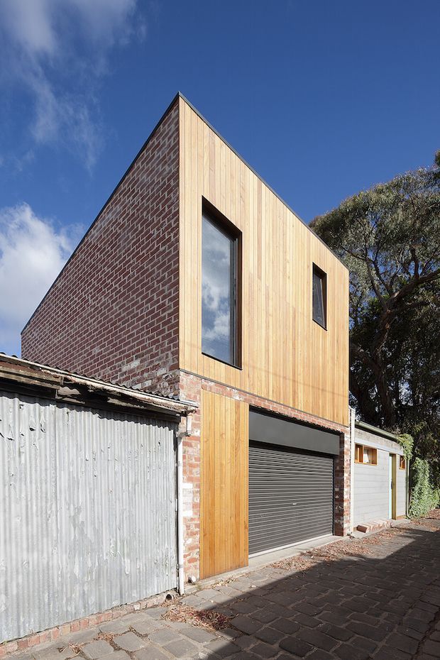 Exterior view of a narrow urban residential extension or garage-front building, combining exposed red brickwork with vertical light timber cladding. The sharply pitched gable form has tall dark-framed windows and a roller garage door at ground level, set along a paved laneway beside mature trees and older structures.