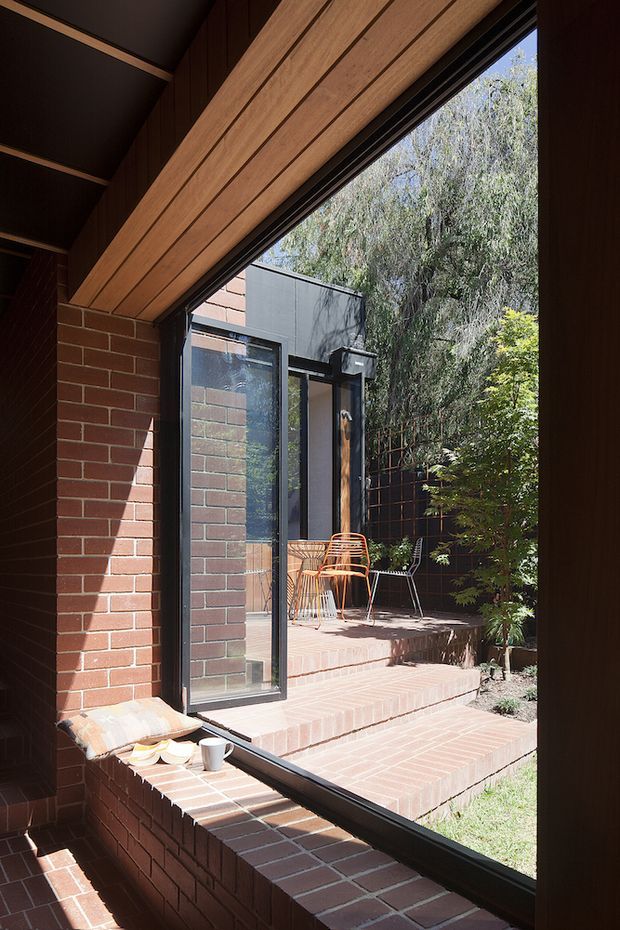 A sunlit interior view looking through a large black-framed glazed opening toward a small brick-paved courtyard or terrace. The room has exposed red brick walls and a dramatic sloped timber-lined ceiling with dark structural framing. Brick steps lead outside to a compact patio furnished with lightweight chairs and a small table, surrounded by dense greenery. The composition highlights the connection between the interior and the private outdoor space.