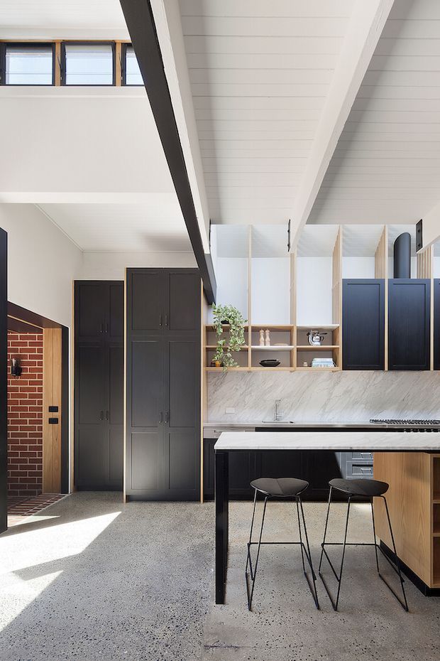 Bright open-plan kitchen and entry area with a high white-lined ceiling and exposed dark structural beam. The kitchen features a large marble-look stone benchtop and splashback, matte black cabinetry, warm timber shelving and island detailing, and two black bar stools. At left is a brick-lined entry opening with tall dark built-in storage. High-level windows and timber-framed glazed partitions bring in natural light.