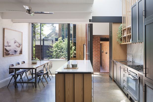 Bright open-plan kitchen and dining area in a renovated contemporary home. The space features a central timber island with a pale stone benchtop, dark blue-grey cabinetry, a stainless steel range cooker and sink, polished concrete flooring, exposed red-brick walls, and large glazed doors opening toward a leafy outdoor area. A dining table sits beside the kitchen, with a ceiling fan and built-in shelving adding functional detail.