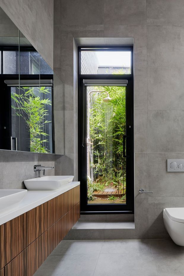 A contemporary bathroom with large grey concrete-look wall and floor tiles, a long timber-veneer floating vanity with two white vessel basins, wall-mounted chrome taps, a frameless mirror, and a white toilet. A tall black-framed glazed door or window opens toward a lush bamboo-filled outdoor area, bringing strong natural greenery into the space.