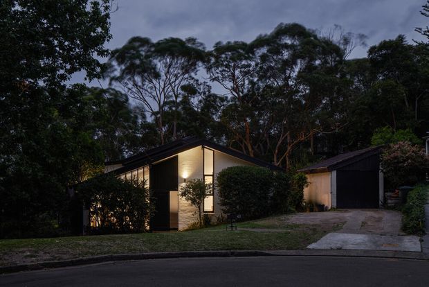Dusk exterior view of a contemporary single-storey residence set among mature trees. The home has a prominent pitched gable roof, dark vertical cladding, warm illuminated windows, a light stone or masonry feature wall, and an attached dark garage reached by a sloped driveway.