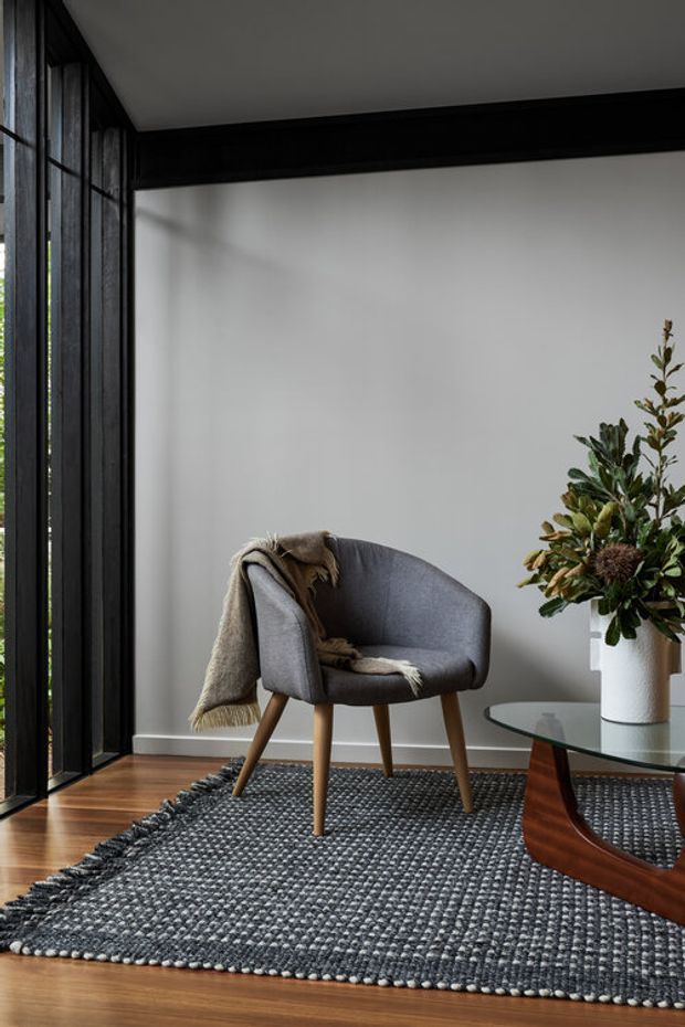 A refined seating corner with a grey upholstered armchair, draped throw, patterned dark area rug, and sculptural wooden glass coffee table. Floor-to-ceiling black-framed glazing and a black overhead structural beam contrast with pale walls and warm timber flooring. A large potted arrangement adds a natural accent.
