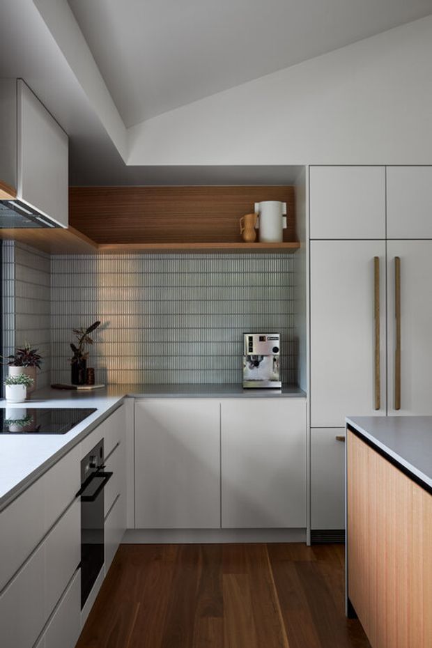 A compact, contemporary kitchen with a U-shaped layout and peninsula. Matte light-grey cabinetry surrounds dark grey benchtops, with long timber handles and a warm timber-clad open shelf above a vertically ridged grey tile splashback. Integrated appliances, an induction cooktop, coffee machine, timber flooring, and a timber-panelled peninsula create a refined, warm modern interior.