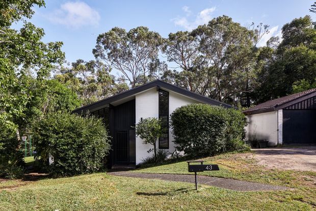Street-level exterior view of a low-rise modern residence with a sharply pitched dark roof, white rendered front wall, dark vertical cladding, and tall narrow glazing. Dense landscaped hedges and mature trees surround the entry, with a paved path and driveway visible. A neighbouring garage or outbuilding appears at right.
