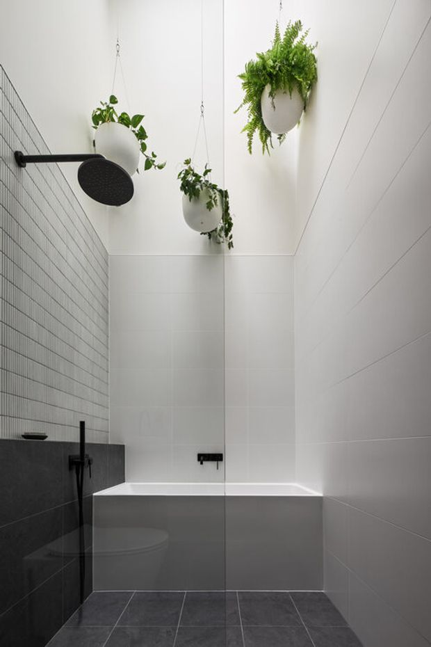 A compact contemporary bathroom with a white built-in bathtub, frameless glass shower screen, black wall-mounted shower fittings, and dark grey tiled flooring and lower wall surfaces. Large white wall tiles create a clean backdrop, while several suspended planters add greenery near the high ceiling. The palette is monochromatic with black accents and natural green plants.