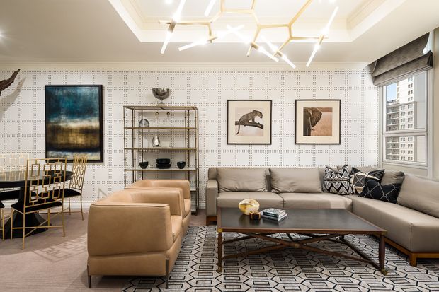 A refined living room with a large sectional sofa, two tan lounge chairs, a dark wood coffee table, patterned rug, open metal shelving, framed artwork, and a small dining or work area. The room features a perforated geometric feature wall, layered tray ceiling, and sculptural contemporary pendant lighting, with high-rise buildings visible through the window.