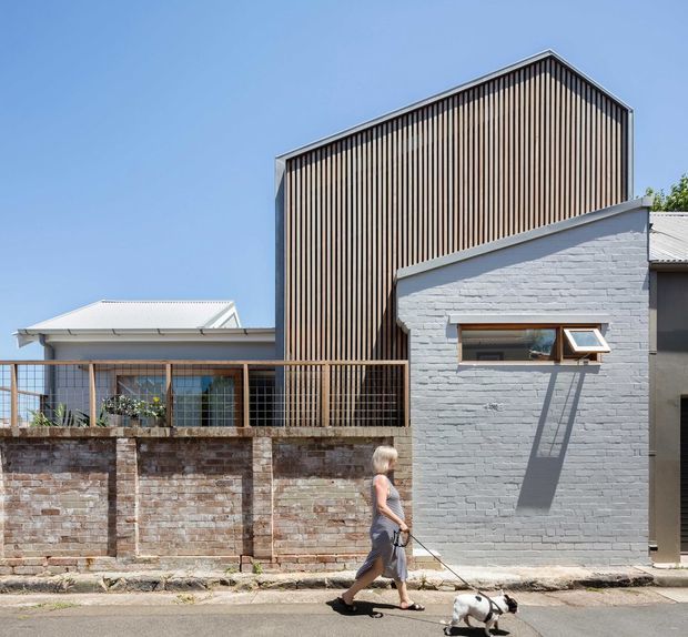 Street-facing view of a contemporary multi-level residential building combining a tall gabled volume clad in closely spaced vertical timber battens with a lower white-painted brick wing. A timber-framed balcony or screened terrace sits above a weathered brick boundary wall, while a small horizontal awning window punctuates the lower facade. Clear blue sky and neighbouring low-rise structures are visible.