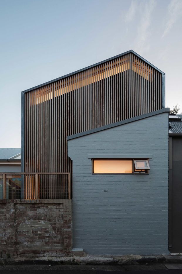 A narrow urban residential facade viewed from the street at dusk. The building combines a dark grey painted brick lower volume with a taller gabled form screened by closely spaced vertical timber battens. Warm interior light glows through the slatted facade and a small horizontal awning window. An aged brick boundary wall and neighbouring rooflines frame the compact infill setting.