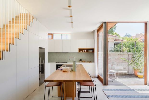 Bright open-plan kitchen and dining area with a light timber island dining table, white handleless cabinetry, pale green textured splashback, integrated appliances, and a sculptural staircase with vertical balustrades. Large timber-framed glazed doors open to a paved courtyard, creating a strong indoor-outdoor connection. The space is clean, contemporary, and softly natural in tone.