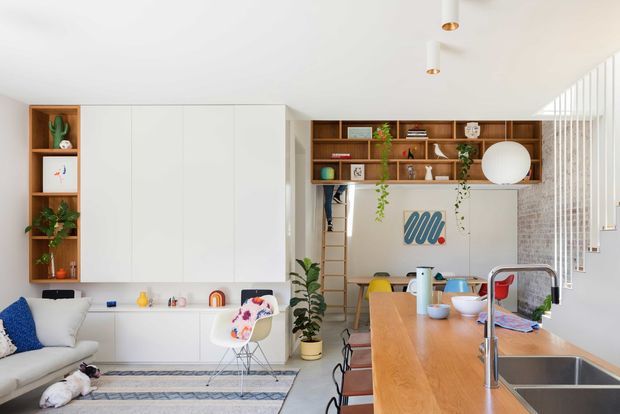 Bright open-plan kitchen and living area with a warm timber island and integrated stainless-steel sink, white full-height cabinetry, open timber shelving, a compact dining setup, and a built-in study nook beneath a loft ladder. The room includes indoor plants, restrained colourful decor, and abundant daylight through vertical glazing or screening.