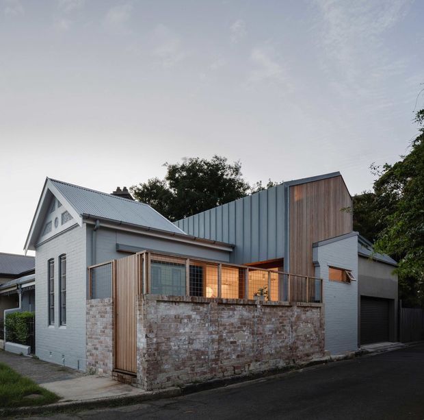 Exterior view of a compact residential extension combining an older gabled brick dwelling with a contemporary addition. The original house has pale painted brick walls, a pitched corrugated metal roof, traditional gable detailing and tall narrow windows. The newer volume uses vertically ribbed blue-grey metal cladding, warm vertical timber screening and extensive glazed openings behind a brick courtyard wall. The home fronts a quiet paved street and is framed by mature vegetation.