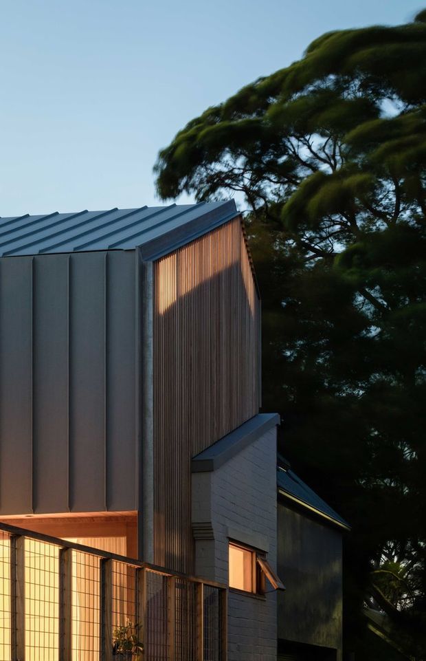 Vertical dusk photograph of a compact contemporary residential building set among mature trees. The facade combines dark standing-seam metal cladding and roofing with a tall finely ribbed timber screen, pale painted masonry, and warm-lit windows. A metal security or privacy fence encloses the lower level, while strong evening light creates a warm contrast against the cool blue-grey exterior and surrounding greenery.