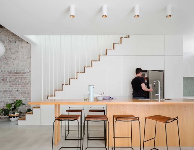Bright open-plan kitchen with a long timber island and integrated seating, white full-height cabinetry, a concealed stairway with timber-edged steps, exposed brickwork, concrete-look flooring, and minimalist cylindrical ceiling lights. A person is using the integrated refrigerator, while bar stools line the island.