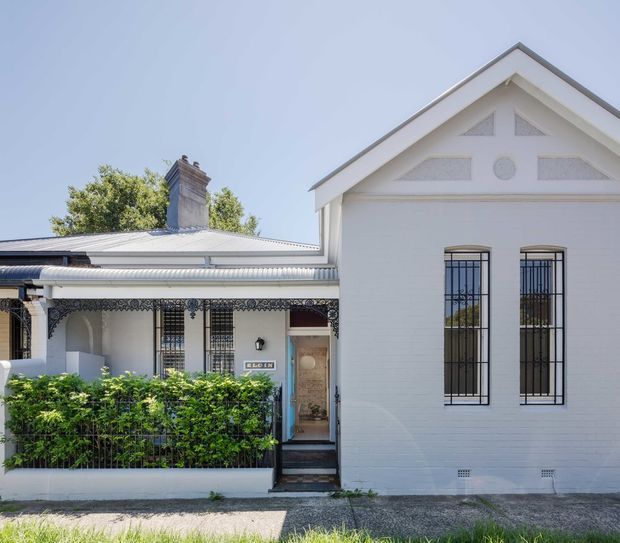 Front exterior view of a renovated heritage-style urban residence, with a white-painted gabled facade, tall barred windows, decorative lacework verandah, covered entry, chimney, and hedge-lined frontage.