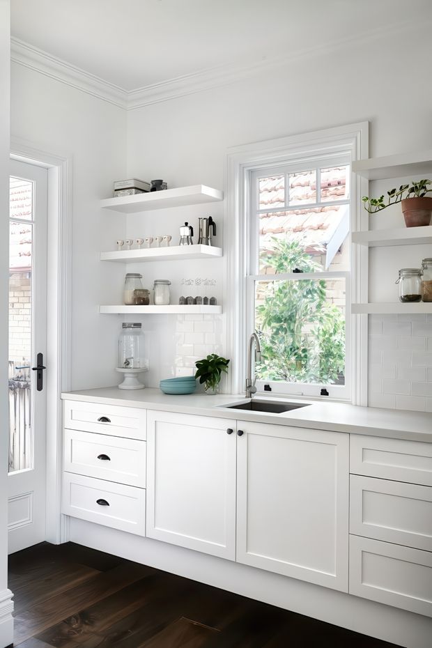 Bright, compact white kitchen with Shaker-style cabinetry, dark timber flooring, white subway-tile splashback, open floating shelves, and a farmhouse-inspired sink beneath a sash window. Traditional moulded cornices and trim frame the space, while small appliances, jars, crockery, and a potted plant add restrained domestic detail.