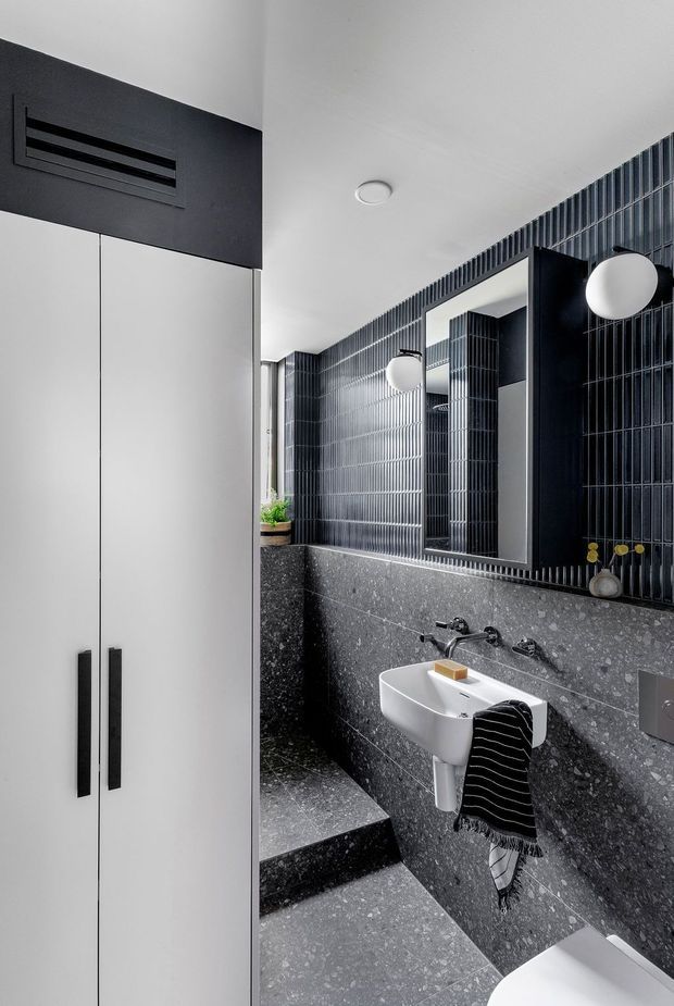 A compact, contemporary bathroom with a monochrome palette. Dark grey terrazzo-style wall and floor surfaces contrast with white cabinetry and a white wall-mounted basin. Glossy vertically ribbed dark tiles form a feature wall around a large rectangular mirror, complemented by black fittings and spherical wall lights. A partial toilet is visible in the foreground, and a step leads toward the vanity area.