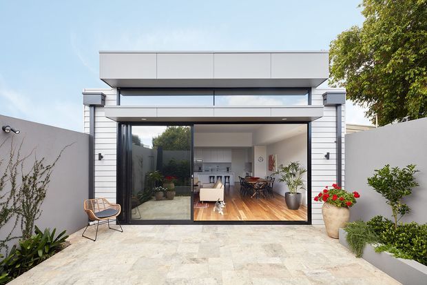 A contemporary single-storey pavilion-style home extension opening onto a paved courtyard. The facade combines horizontal light cladding, dark-framed floor-to-ceiling sliding glazing, a deep projecting roof canopy, and a clean parapet-like upper form. Inside, a bright open-plan living and dining area with timber flooring is visible, while the courtyard includes restrained planting and a simple outdoor chair.