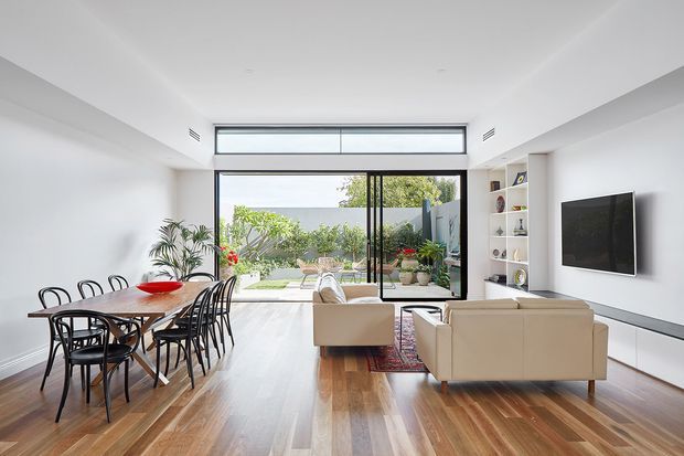 Bright open-plan interior combining a dining area and living room, with white walls, built-in shelving, a wall-mounted television, large glazed doors and clerestory windows opening to a landscaped courtyard. The room has warm timber flooring, a neutral sofa arrangement, black dining furniture, and abundant natural light.