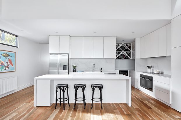 Bright contemporary white kitchen with a large central island and three black bar stools. The room features extensive white flat-panel cabinetry, integrated stainless-steel refrigerator, built-in oven and microwave, white benchtops and splashback, a small sink, and a built-in wine rack. Warm timber flooring contrasts with the white cabinetry, while a framed artwork and radiator sit along the left wall.