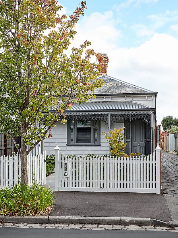 Front exterior view of a compact historic single-storey cottage, featuring a pale rendered facade, ornate decorative verandah and lacework trim, grey timber shutters and entry door, a pitched slate-look roof with brick chimney, and a white picket fence. A paved driveway and established garden planting frame the street-facing facade.