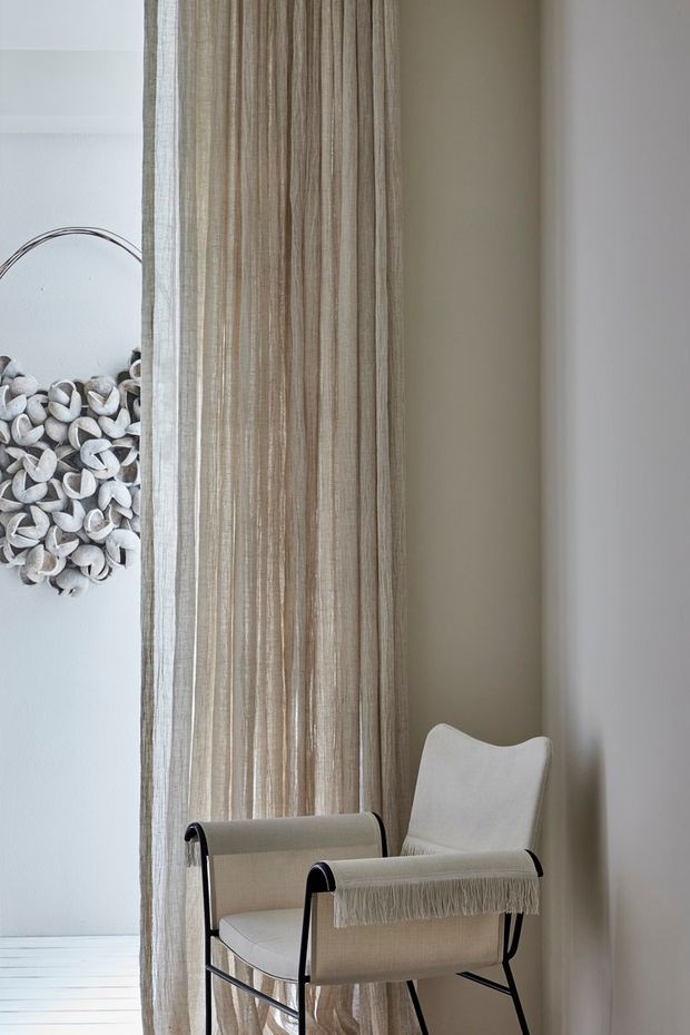 A minimalist, softly lit interior corner featuring a neutral-toned upholstered lounge chair with a black metal frame positioned beside floor-to-ceiling sheer curtains. White walls and pale flooring create an airy, understated setting, with a small shell wall decoration visible at the edge.