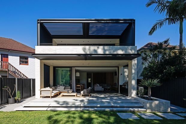 Exterior rear view of a contemporary multi-level home with a projecting upper pavilion, open slatted roof structure, white brick facade, broad covered terrace, large glazed living area, and landscaped lawn with palms. The image highlights the building envelope, balcony/terrace, and indoor-outdoor connection.