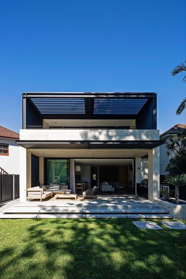 Front exterior view of a contemporary two-storey residence with a crisp white brick facade, broad recessed ground-floor opening, and a large upper-level outdoor room shaded by a black louvred pergola. Pale stepped paving leads from the lawn to an outdoor seating area, with glimpses of the living room and dark built-in joinery visible inside. Strong sunlight creates tree shadows across the facade and terrace.